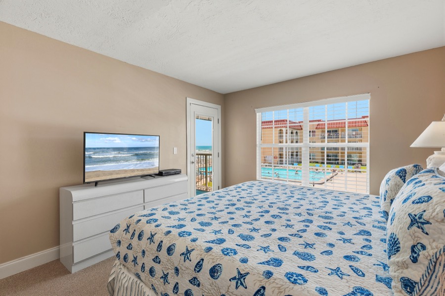 Primary bedroom with seashell quilt, TV, and windows framing pool and ocean views